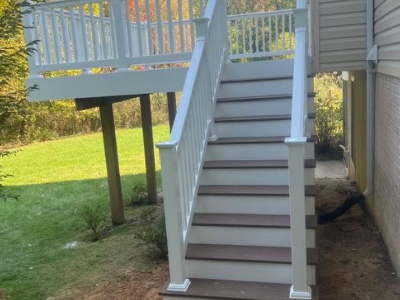 White wooden staircase with railings leads up to an elevated deck attached to a house, in a sunny backyard with autumn trees in the background.